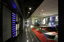 Server racks line a dark hallway next to a glass-walled office with desks and computers.