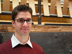 A man with dark hair and glasses wearing a maroon sweater and collared shirt.