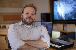 A bearded man in a light blue shirt smiles with arms crossed in a music production studio.