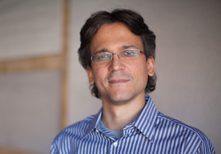 A man with dark wavy hair and glasses wearing a blue and white striped button-down shirt.