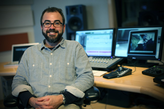 A man with glasses and a beard sits in front of computer monitors and audio equipment in a studio.
