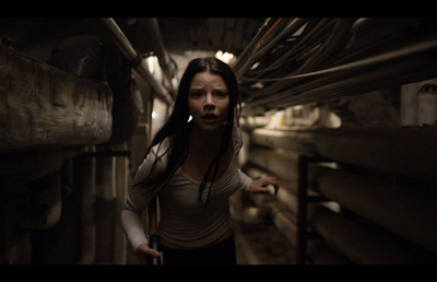 A young woman with long dark hair looks around with a frightened expression in a dark, narrow industrial hallway.