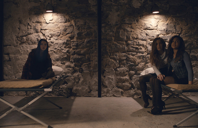 Three women sit on cots in a dimly lit room with rough stone walls and overhead lights.