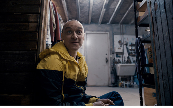 A bald man in a yellow and black jacket sits on a basement floor leaning against a wooden wall.