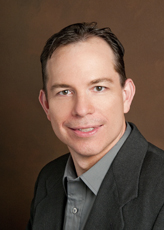 A man with short dark hair wearing a grey collared shirt and a dark blazer against a brown background.