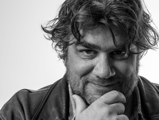 Black and white close-up portrait of a man with wavy hair resting his chin on his hand.