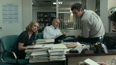 Four people in an office look at documents on a desk piled high with stacks of paper.