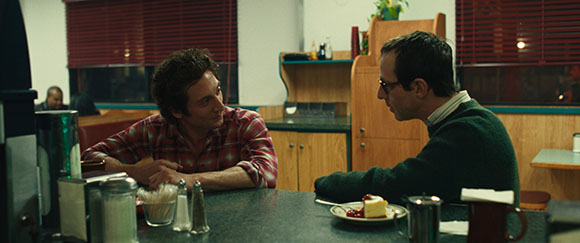 Two men sit across from each other at a dark diner counter with a slice of cheesecake on the table.