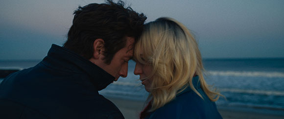 A man and a blonde woman lean their foreheads together against a dark ocean background at dusk.