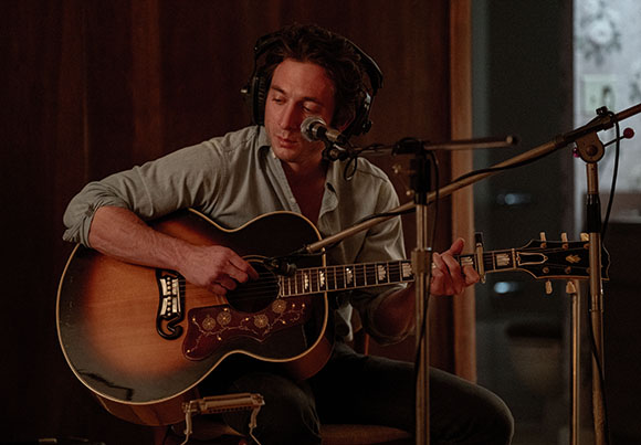 A man in a light shirt plays an acoustic guitar and sings into a microphone while wearing headphones.
