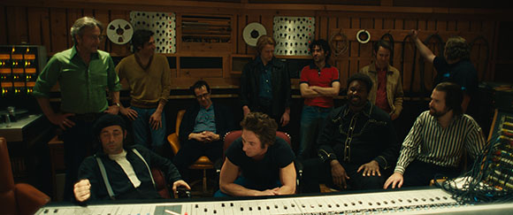 A group of men sitting and standing around a large mixing console in a wood-paneled recording studio.