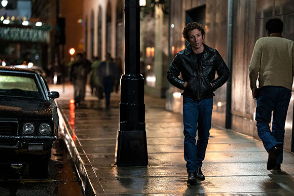 A man in a black leather jacket and blue jeans walks along a wet city sidewalk at night.