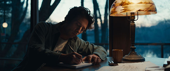 A man sits at a table writing in a notebook by the light of a shaded lamp near a window at dusk.