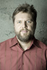A man with brown hair and a beard wearing a red button-down shirt with vertical stripes.