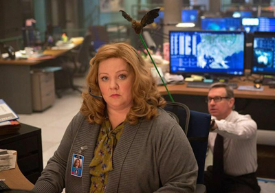 A woman with wavy blonde hair wearing a grey cardigan sits in a busy office with computer monitors.