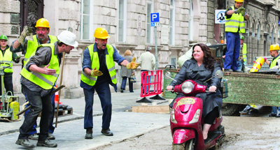 Construction workers in yellow vests gesture toward a woman riding a pink scooter past a work site.
