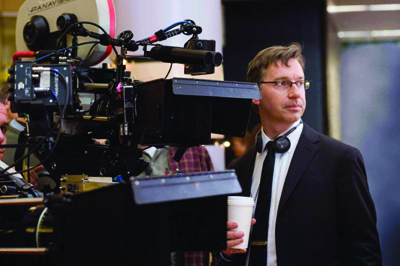 A man in a dark suit and glasses stands beside a large professional motion picture camera.
