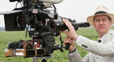A man in a tan hat and suit jacket adjusting a large professional film camera in a field.