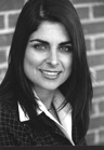 A smiling woman with long dark hair wearing a collared shirt and patterned jacket.