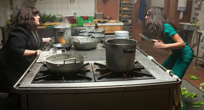 Two women stand on opposite sides of a large stove with several metal pots.