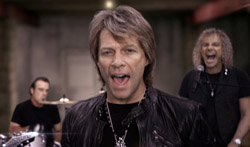 A man in a black leather jacket sings while a drummer and another man stand behind him.