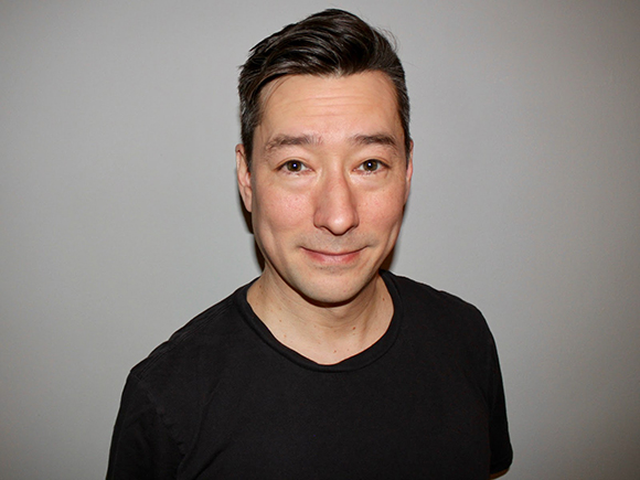 A man with short dark hair and a black t-shirt looking forward against a plain gray background.