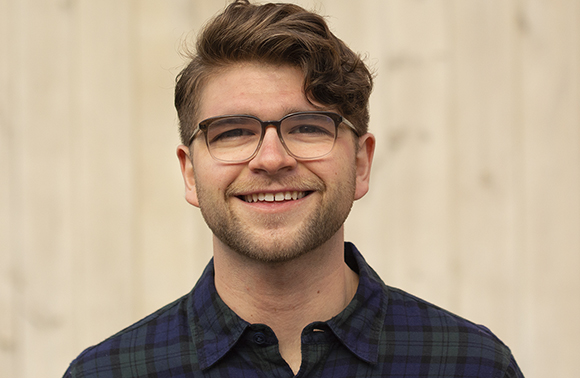 A man with short brown hair and glasses smiling against a light-colored background.