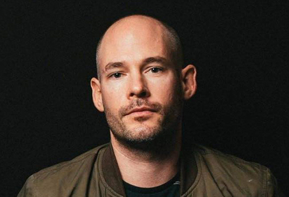 A bald man with light facial hair looking directly at the camera against a black background.