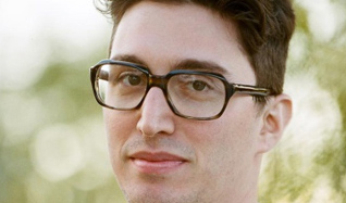 A man with dark hair and dark-rimmed glasses looking forward against a blurred outdoor background.