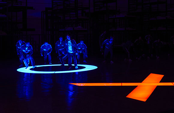 Performers in blue jumpsuits stand on a dark stage illuminated by blue and orange light projections.