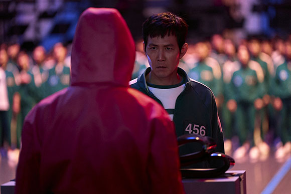 A man in a green tracksuit labeled 456 stares intensely at a person in a red hooded jumpsuit.