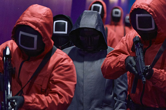 People in red and grey hooded jumpsuits with black masks featuring white geometric shapes.
