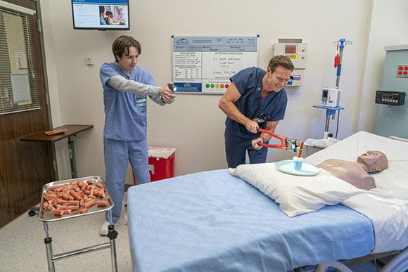 Two people in scrubs performing a medical procedure on a training mannequin in a classroom.