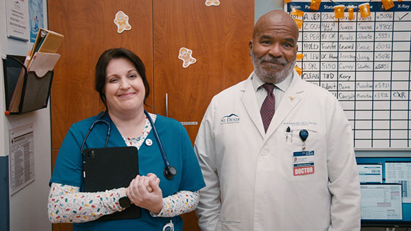 A man in a white lab coat and a woman in teal scrubs stand in a medical office.