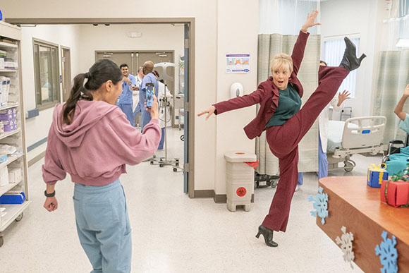 A woman in a maroon suit performs a high kick in a hospital hallway near medical staff.