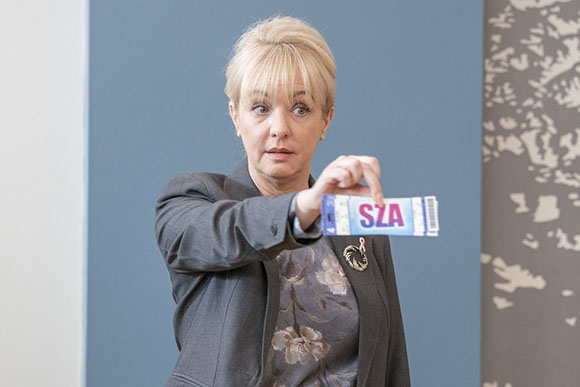 A blonde woman in a grey blazer holds out a small blue and white packet with the letters SZA on it.
