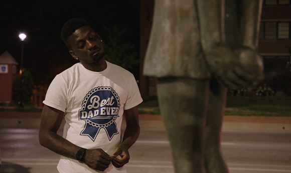 A man in a white 'Best Dad Ever' t-shirt looks down while standing next to a large statue at night.