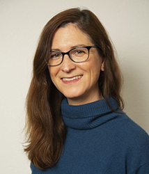 A smiling woman with long brown hair and black-rimmed glasses wearing a blue turtleneck.