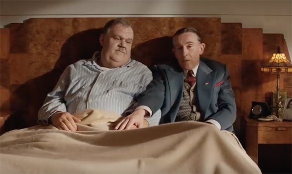 A man in striped pajamas and a man in a suit sit together in a bed under a tan blanket.