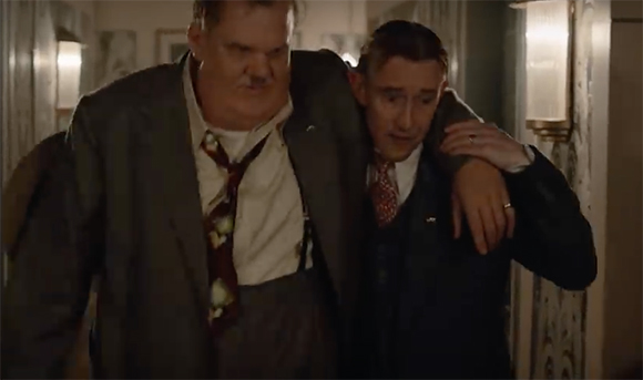 A large man in a grey suit supports a slimmer man in a dark suit as they walk through a hallway.