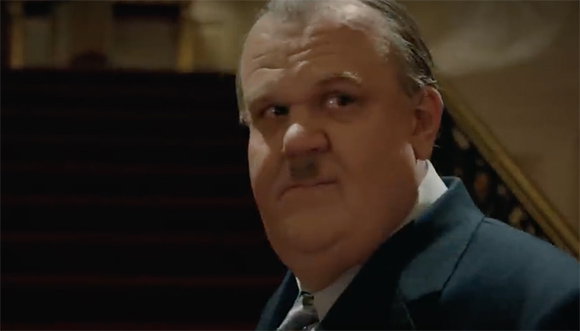 A middle-aged man with a mustache and receding hairline looks sideways in a dimly lit room.