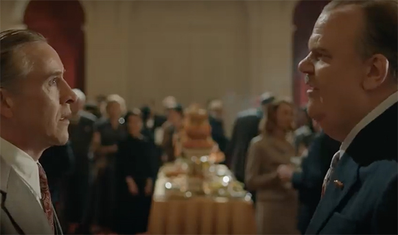 Two men in formal attire engage in conversation in a large, dimly lit ballroom filled with people.