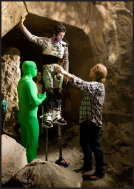 A person in a patterned costume on tall stilts stands in a stone structure while others assist them.