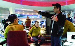 A man in a black jacket and cap gestures toward actors on a futuristic spaceship set.