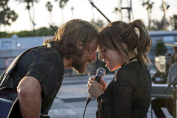 A man with long hair and a woman with a ponytail lean their foreheads together outdoors.