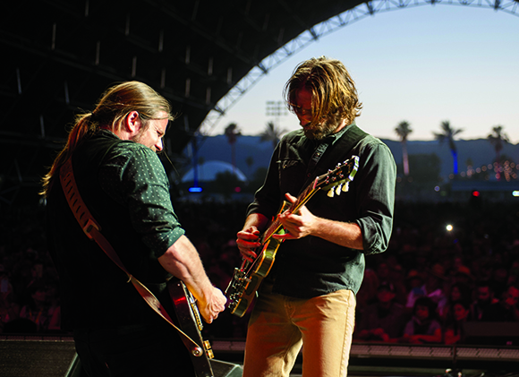 Two men playing electric guitars on a stage during an outdoor evening concert.