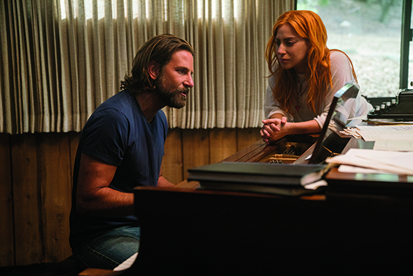 A bearded man in a blue shirt sits at a piano, looking up at a red-haired woman leaning on the instrument.