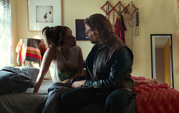 A man in a leather jacket and a woman in a white tank top sit facing each other on a bed.
