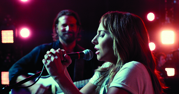 A woman sings into a microphone while a man plays guitar under red and white stage lights.