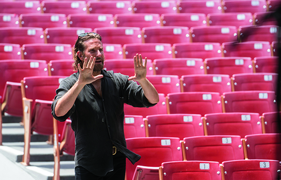 A man with long hair and a beard gestures with open palms in front of rows of empty red seats.